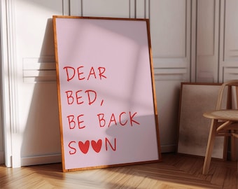 May include: A framed pink print with the text "DEAR BED, BE BACK SOON" in red. Two red hearts are below the text. The frame is brown, and the print is leaning against a white wall. The floor is made of wood.