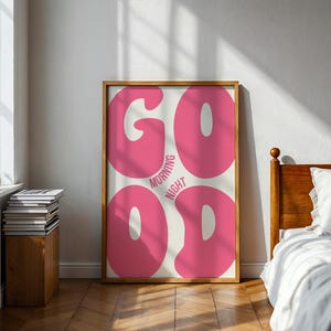 Może przedstawiać: Oprawiony plakat z napisem "GOOD MORNING NIGHT" dużymi, r&oacute;żowymi literami w stylu bąbelkowym. Plakat jest w drewnianej ramie i opiera się o białą ścianę. Pok&oacute;j ma drewnianą podłogę i ł&oacute;żko.