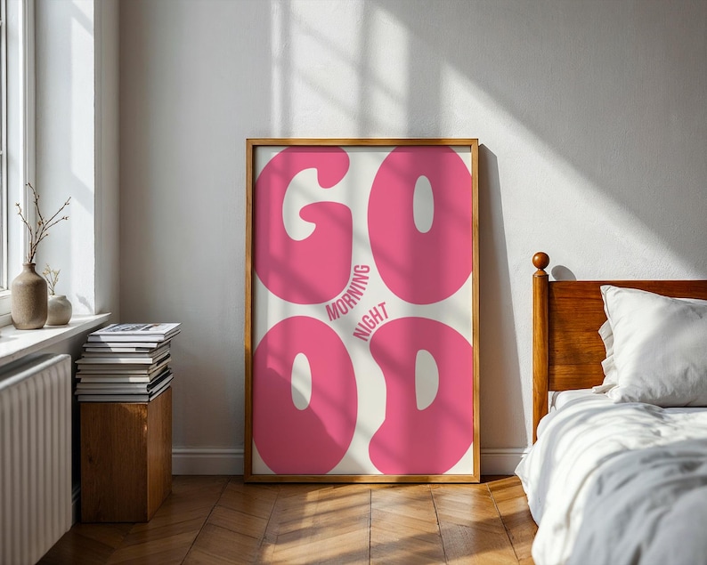 Może przedstawiać: Oprawiony plakat z napisem "GOOD MORNING NIGHT" dużymi, r&oacute;żowymi literami w stylu bąbelkowym. Plakat jest w drewnianej ramie i opiera się o białą ścianę. Pok&oacute;j ma drewnianą podłogę i ł&oacute;żko.
