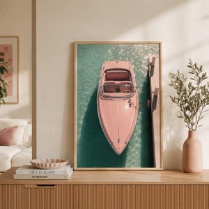 May include: A framed vertical wall art print featuring a top-down view of a pastel pink motorboat docked alongside a wooden pier in clear turquoise water. The boat has a windshield and bench seating, captured in a bright, sunlit outdoor setting.