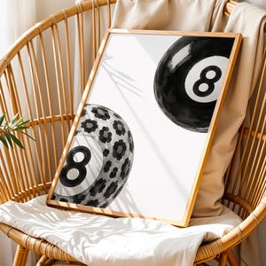 May include: A rectangular framed print featuring two black and white eight balls on a white background. One ball is solid black with a white number eight, while the other ball is patterned with black and gray floral shapes and a white number eight.
