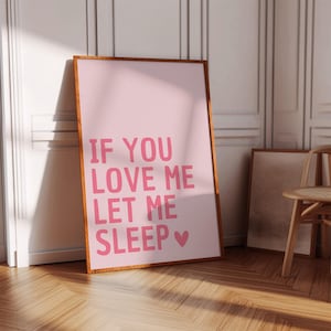 May include: A framed pink print with the text "IF YOU LOVE ME LET ME SLEEP" in pink letters. The print is in a wooden frame and leans against a white wall. A wooden chair and another framed print are in the background.