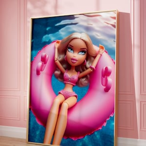 以下が含まれることがあります： ピンクのビキニと浮き輪を着けた人形がプールにいるフレームプリント。人形はブロンドの髪と青い目です。プリントは金色のフレームが付いたピンクの壁に表示されています。