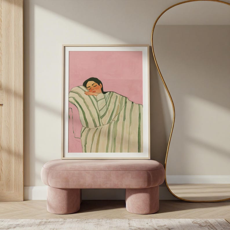 May include: A framed art print featuring a person resting on a sofa, draped in a green and white striped blanket. The artwork has a pink background and is displayed above a pink, rounded ottoman. A gold-framed mirror is visible.
