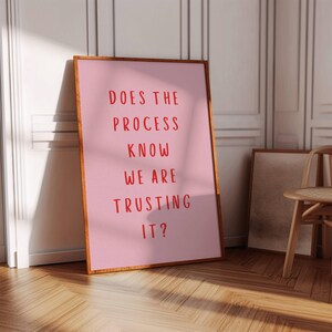 Op de afbeelding: Een roze ingelijste poster met de tekst "DOES THE PROCESS KNOW WE ARE TRUSTING IT?" in rode letters.