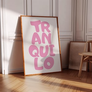 May include: A framed art print with the word "TRANQUILO" in large, bubble-style pink letters. The print has a white background and a wooden frame. The artwork is leaning against a wall in a room with wooden floors.