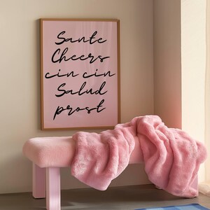 Puede incluir: Un banco rosa con una manta rosa esponjosa. Encima, un estampado rosa enmarcado muestra las palabras "Sante, Cheers, cin cin, Salud, prost" en escritura negra. La escena es interior.