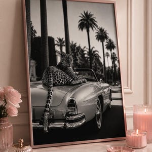 May include: A framed black and white wall art print featuring a leopard resting on the trunk of a vintage convertible car. The background displays a street lined with tall palm trees. The frame is a metallic rose gold color, presented on a surface.
