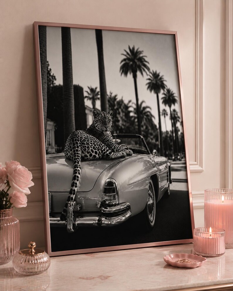May include: A framed black and white wall art print featuring a leopard resting on the trunk of a vintage convertible car. The background displays a street lined with tall palm trees. The frame is a metallic rose gold color, presented on a surface.