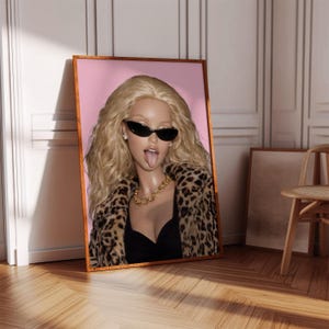 May include: Framed art print featuring a blonde woman wearing black sunglasses, a black top, and a leopard print coat. The woman is sticking out her tongue. A gold chain necklace is visible. The background is pink.