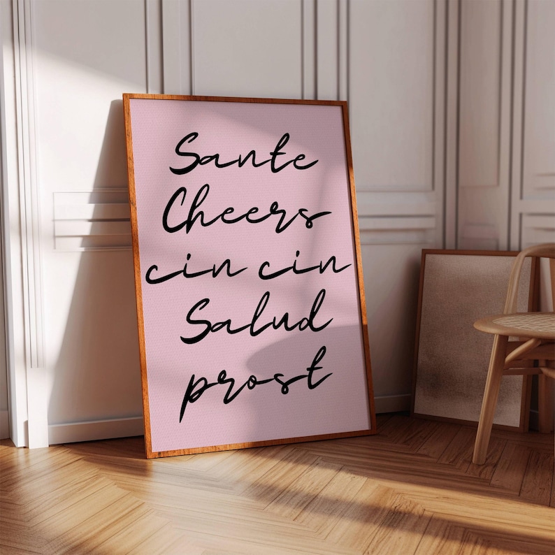 Puede incluir: Un estampado rosa enmarcado con las palabras "Sante, Cheers, cin cin, Salud, prost" en escritura negra. El marco es de color marr&oacute;n c&aacute;lido, y el estampado est&aacute; apoyado contra una pared blanca y un suelo de madera.