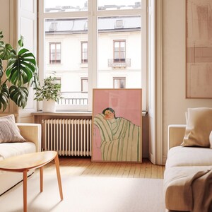 May include: A framed art print featuring a person resting on a striped blanket. The artwork has a pink background and a wooden frame. The room has a window, a sofa, and a wooden coffee table.