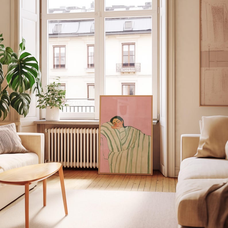 May include: A framed art print featuring a person resting on a striped blanket. The artwork has a pink background and a wooden frame. The room has a window, a sofa, and a wooden coffee table.