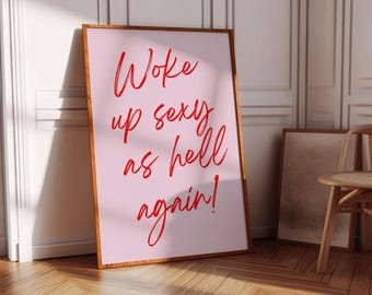 May include: A framed art print with the text "Woke up sexy as hell again!" in red script on a pink background. The frame is a warm brown color, and the print is leaning against a white wall and wooden floor.