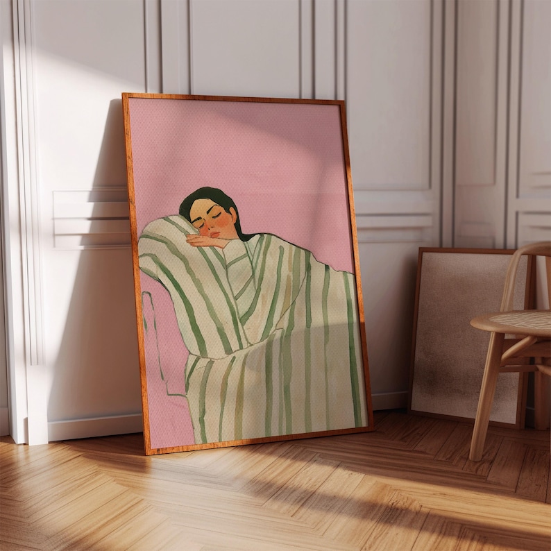 May include: A framed art print featuring a person resting on a pink surface. The person is wearing a green and white striped garment. The artwork is in a wooden frame and leans against a white wall.