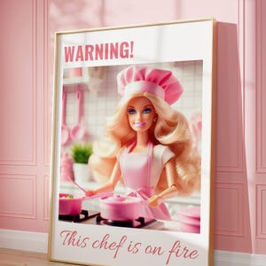 Peut inclure: Une impression encadrée avec un fond rose et une bordure blanche. L'impression représente une poupée Barbie habillée en chef, tenant une casserole rose et une spatule rose. Le texte "ATTENTION ! Ce chef est en feu" est imprimé sous l'image.