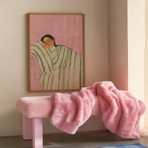 May include: A pink bench with a plush, fuzzy texture is draped with a matching pink blanket. Above, a framed artwork depicts a person resting, wrapped in a striped blanket. The room has a soft, pastel color palette.