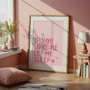 May include: A framed pink print with the text "IF YOU LOVE ME LET ME SLEEP" in a bold, pink font. The print is displayed in a room with a pink wall, a window, and a wooden floor. A pink pillow and a lamp are also visible.