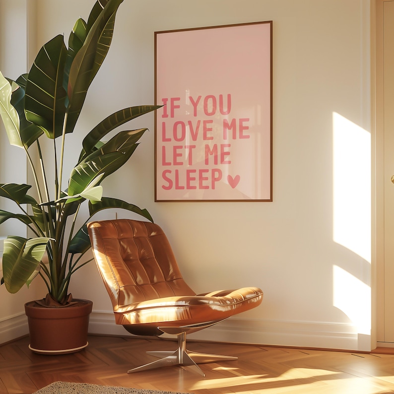 May include: A framed pink poster with the text "IF YOU LOVE ME LET ME SLEEP" hangs on a white wall. A brown leather chair sits in front of the wall, and a large potted plant is to the left. The room has a warm, inviting atmosphere.