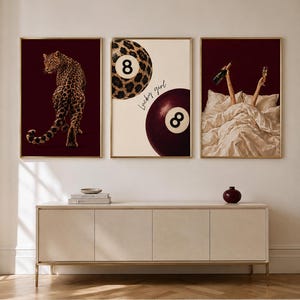 Op de afbeelding: Drie ingelijste wandposters boven een dressoir. De linker poster toont een luipaard op een donkerrode achtergrond. De middelste poster bevat twee achtballen, één met een luipaardprint, en de tekst "Lucky girl". De rechter poster toont armen die een fles en glas in bed vasthouden.