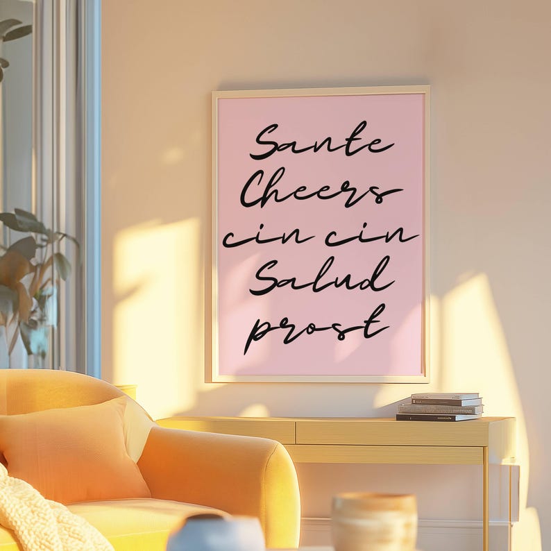Puede incluir: Un estampado rosa enmarcado con texto negro en cursiva que dice "Sante, Cheers, Cin Cin, Salud, Prost". El estampado se muestra en una pared encima de una mesa de madera. Un sill&oacute;n naranja est&aacute; en primer plano.