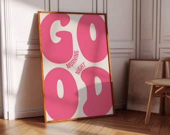 Op de afbeelding: Een ingelijste kunstdruk met het woord "GOOD" in grote, roze letters in bubbelstijl, met "MORNING" en "NIGHT" in kleinere tekst. De print heeft een crèmekleurige achtergrond en een houten lijst. Het kunstwerk leunt tegen een muur.