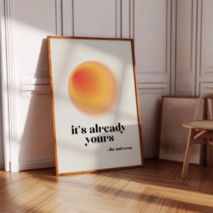 May include: A framed art print with a wooden frame. The print features a blurred orange and yellow orb against a white background. The text "it's already yours - the universe" is printed in black at the bottom of the print. The print is leaning against a wall.