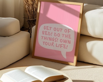 May include: A framed pink print with the text "GET OUT OF BED! DO THE THINGS! OWN YOUR LIFE!" in a white speech bubble. An open book rests on a round table in front of the print.