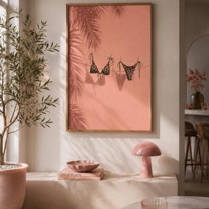 May include: A framed wall art print featuring a coral-colored background with a clothesline displaying a leopard print bikini top and bottom, accompanied by the shadows of palm fronds cast across the surface of the artwork.