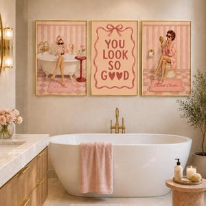 May include: A set of three framed wall art prints featuring pink and white striped backgrounds. The left print shows a person in a bathtub, the center print displays the text YOU LOOK SO GOOD with a bow, and the right print shows a person with a mirror.