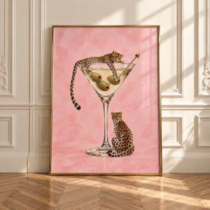 May include: A framed wall art print featuring a large martini glass filled with liquid and three green olives on a solid pink background. One leopard rests its body across the rim of the glass, while a second leopard sits at the base.