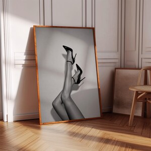 May include: A framed black and white wall art print featuring a pair of legs wearing black high-heeled stiletto shoes with ankle straps. The artwork is displayed in a wooden frame, showcasing a monochromatic photographic composition of footwear.