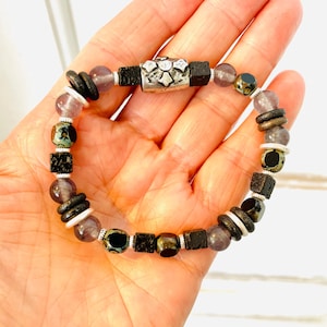 May include: A beaded bracelet with a silver charm, featuring black, brown, and purple beads. The bracelet is made with natural materials and has a rustic, earthy aesthetic.
