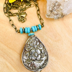 large tibetan repousse pendant necklace. The pendant has an intricate floral design called repousse and is attached to a brass chain with turquoise beads.