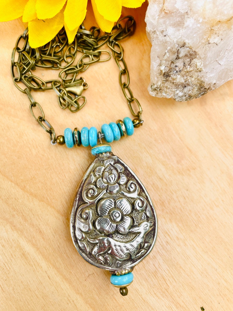large tibetan repousse pendant necklace. The pendant has an intricate floral design called repousse and is attached to a brass chain with turquoise beads.