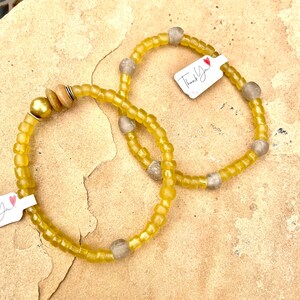 May include: Two yellow beaded bracelets with gray beads and wooden accents. One bracelet has a gold bead and a tag that says "Thank You!" in black text. The bracelets are on a textured, tan surface.