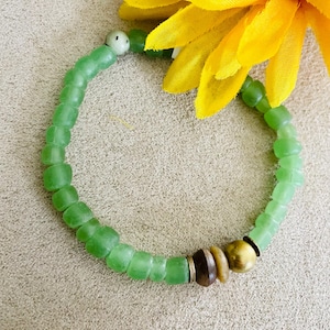 May include: A green beaded bracelet with a mix of square and round beads. The bracelet features a variety of natural-toned beads, including brown and gold, and is accented with a small, white, round bead. A yellow flower is in the background.