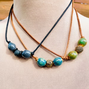 May include: A layered necklace with brown leather cord and black cord. The necklace features blue and green ceramic beads and a gold metal bead.