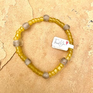 May include: A yellow beaded bracelet with six gray beads. The bracelet is on a light brown surface. A white tag with the words "Thank You" and a red heart is attached to the bracelet.