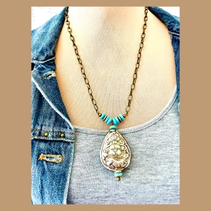 large tibetan repousse pendant necklace. The pendant has an intricate floral design called repousse and is attached to a brass chain with turquoise beads.