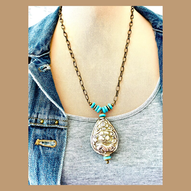 large tibetan repousse pendant necklace. The pendant has an intricate floral design called repousse and is attached to a brass chain with turquoise beads.