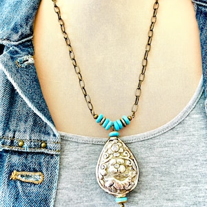 large tibetan repousse pendant necklace. The pendant has an intricate floral design called repousse and is attached to a brass chain with turquoise beads.