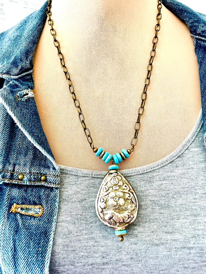 large tibetan repousse pendant necklace. The pendant has an intricate floral design called repousse and is attached to a brass chain with turquoise beads.
