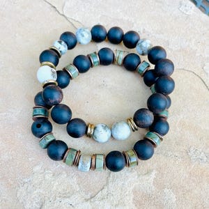 May include: Two beaded bracelets. Each bracelet features dark blue matte beads, interspersed with white and gray speckled beads, gold-colored spacers, and green and bronze-colored cylindrical beads. The bracelets are stacked on a light brown surface.