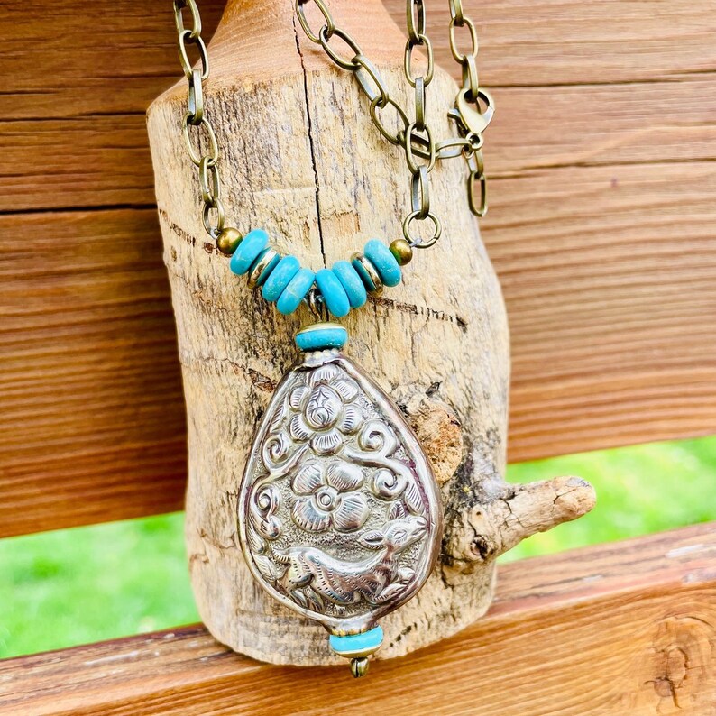 large tibetan repousse pendant necklace. The pendant has an intricate floral design called repousse and is attached to a brass chain with turquoise beads.