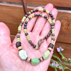 May include: A handmade necklace featuring a mix of green, brown, and cream-colored beads. The necklace has a variety of bead shapes, including small round beads and larger, faceted stones. The clasp is a bronze-colored lobster claw.