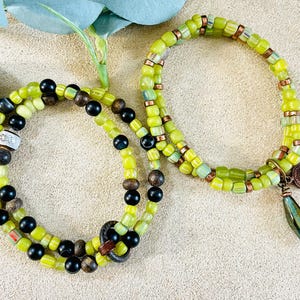 May include: Two beaded bracelets. One bracelet features olive green and black beads, with a few brown beads. The other bracelet has olive green beads, copper-colored spacers, and a decorative charm with a green pendant.