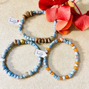May include: Three beaded bracelets with blue, white, and orange beads. One bracelet has a gold bead and brown beads. Each bracelet has a small tag that says "Thank You". A red flower is in the background.