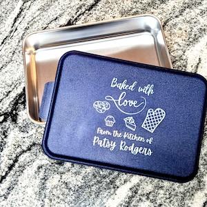 May include: A rectangular metal baking pan with a dark blue lid. The lid features white text that reads Baked with love from the Kitchen of Patsy Rodgens, accompanied by white line art icons of cookies, a cupcake, a slice of cake, and an oven mitt.
