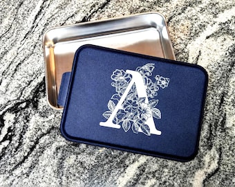 Könnte beinhalten: Ein rechteckiger Aufbewahrungsbehälter aus Metall mit abnehmbarem Deckel. Der Deckel ist dunkelblau und zeigt einen großen weißen Buchstaben A, der von einem detaillierten weißen Blumenmuster umgeben ist. Das Innere ist aus poliertem Silbermetall.